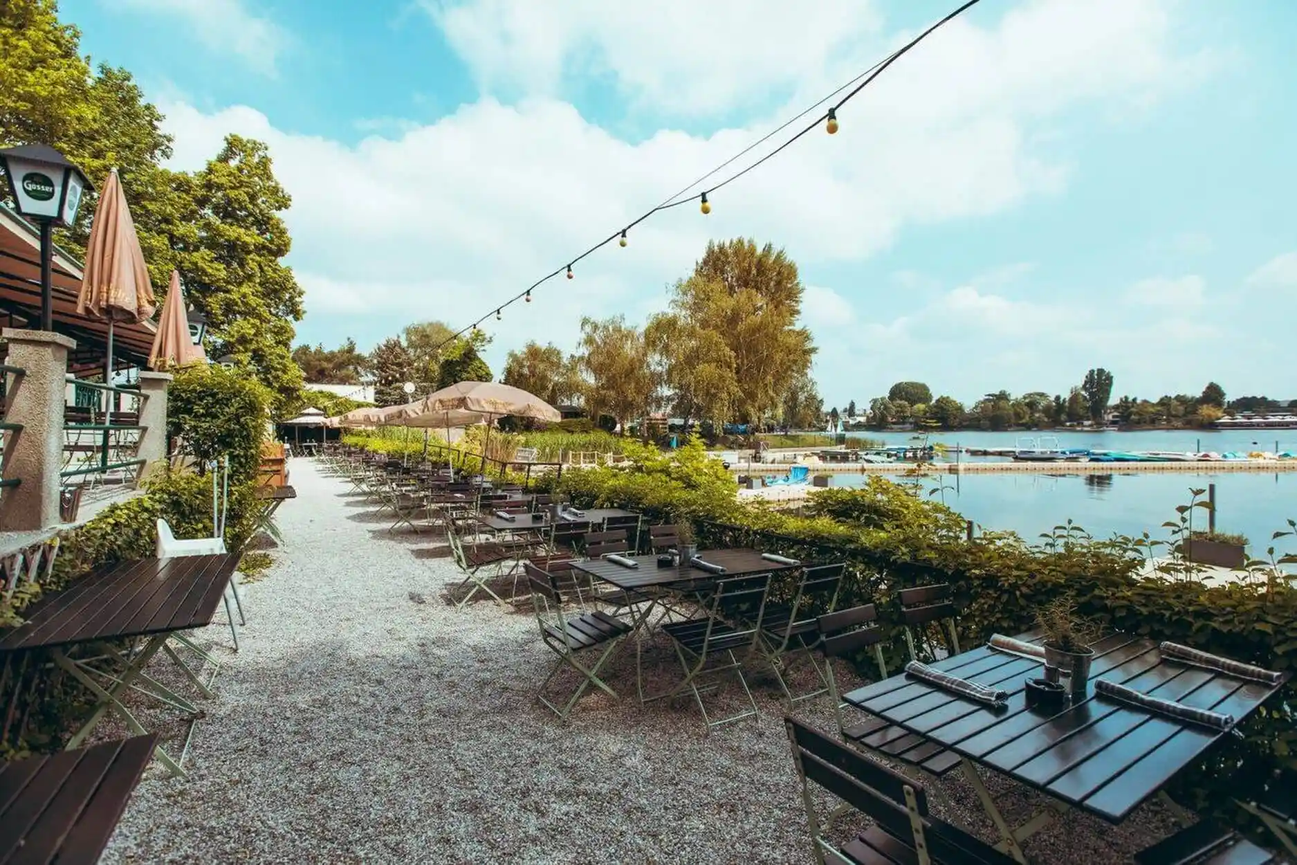 Ein Café im Freien am See mit leeren Tischen und Stühlen auf einem Kiesweg, Lichterketten über dem Kopf, üppigem Grün und Menschen, die im See schwimmen, unter einem blauen Himmel mit vereinzelten Wolken.