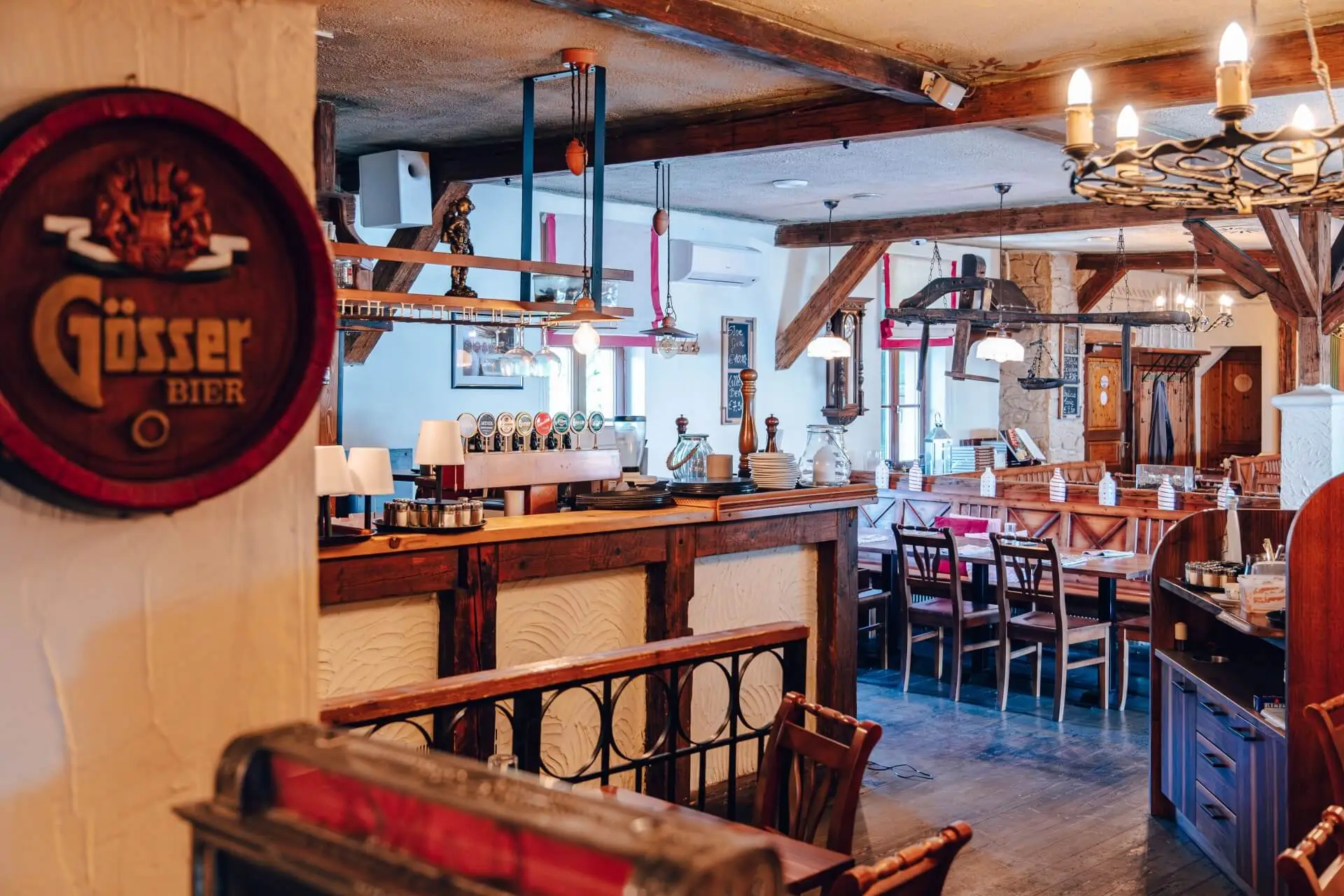 Gemütliches Restaurantinterieur mit Holzbalken, Tischen und Stühlen, rustikalem Dekor, Hängelampen und einem Gössser Bier-Schild an der Wand. Der Raum wirkt warm und einladend, mit einer Bar und einem sichtbaren Essbereich.