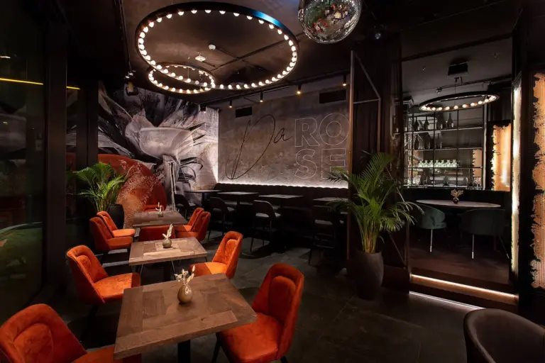 Eine stilvolle, schwach beleuchtete Lounge mit orangefarbenen Samtstühlen, dunklen Holztischen, modernen runden Leuchten, Pflanzen, einem Wandgemälde mit der Aufschrift La Rose und einer gut gefüllten Bar im Hintergrund.