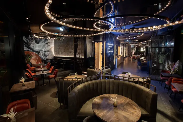 Ein modernes, schummriges Restaurant mit geschwungenen Ständen, runden Holztischen und dramatischen, von der Decke hängenden runden Leuchten. Die Einrichtung ist in dunklen Tönen gehalten, die Sitze sind weich und die Wände kunstvoll gestaltet.