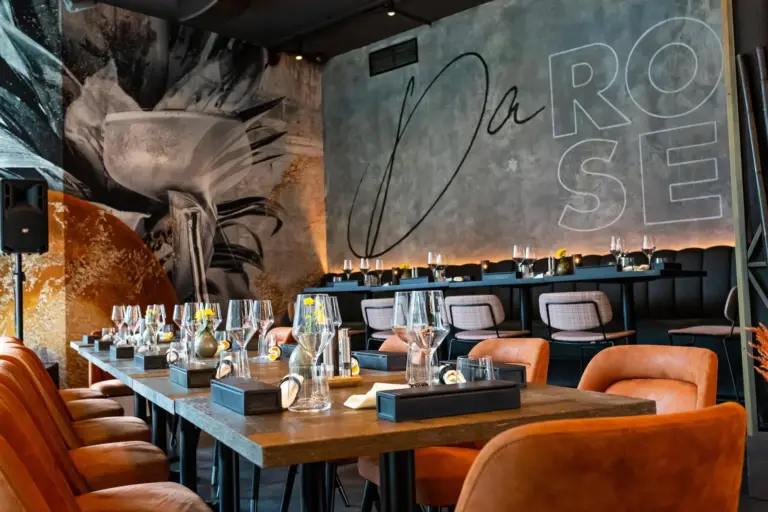 Ein stilvolles, modernes Restaurant mit orangefarbenen Ledersesseln, Holztischen mit Gläsern und gelben Blumen und einem großen Wandgemälde mit dem Schriftzug "Da ROSE" in fetten Buchstaben.