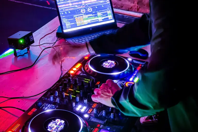 Ein DJ bedient ein farbenfrohes, beleuchtetes Mischpult neben einem Laptop, auf dem eine Musiksoftware angezeigt wird, mit leuchtender rosa und blauer Beleuchtung in einem Nachtclub oder auf einer Party.