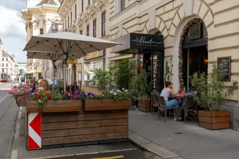 Eine Straßenansicht eines europäischen Cafés mit Sitzgelegenheiten im Freien, Topfpflanzen und Blumen. Die Leute sitzen an Tischen unter einem großen Sonnenschirm. Das Café befindet sich im Erdgeschoss eines historischen Gebäudes.