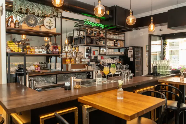 Eine gemütliche Bar mit Holztischen und schwarzen Stühlen und einer gut bestückten Theke mit Flaschen, Gläsern und Fassbierzapfanlagen. Über dem Kopf hängen warme Hängelampen, und im Hintergrund ist ein grünes Tanqueray-Schild zu sehen.