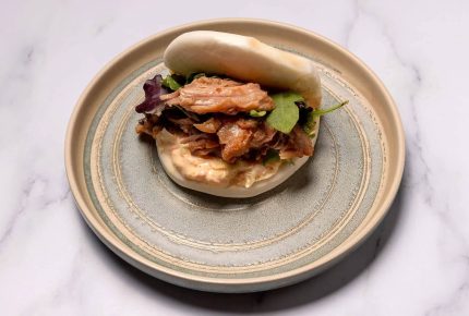 Ein gedämpftes Bao-Brötchen, gefüllt mit zerkleinertem Schweinefleisch, Blattgemüse und cremiger Sauce, serviert auf einem beigen Teller mit blau-grauem Rand auf Joyces Marmoroberfläche.