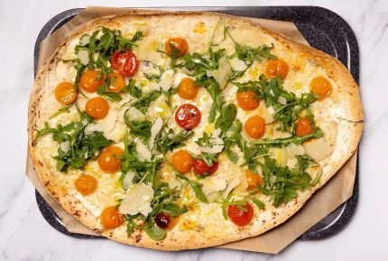 Ovale Fladenbrotpizza, belegt mit geschmolzenem Käse, gelben und roten Kirschtomaten, frischem Rucola und gehobeltem Parmesan - die typische Note von Joyce -, serviert auf braunem Pergamentpapier auf einem dunklen Backblech.