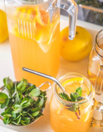 Ein Krug und ein Einmachglas mit geeistem Zitronendrink mit Zitronenscheiben und Minze, daneben eine Schale mit frischem Basilikum und eine ganze Zitrone auf einem weißen Tablett. Das Glas, garniert mit Minze und einem Joyce-Metallstrohhalm, sieht sehr erfrischend aus.