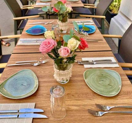 Ein langer Esstisch im Freien, gedeckt für eine Mahlzeit mit grünen und blauen Tellern, Besteck, Gläsern und floralen Tafelaufsätzen mit rosa und weißen Rosen in Gläsern. Schwarze Stühle stehen auf beiden Seiten des Tisches und schaffen eine Szene, die an ein Treffen im Garten von Joyce erinnert.