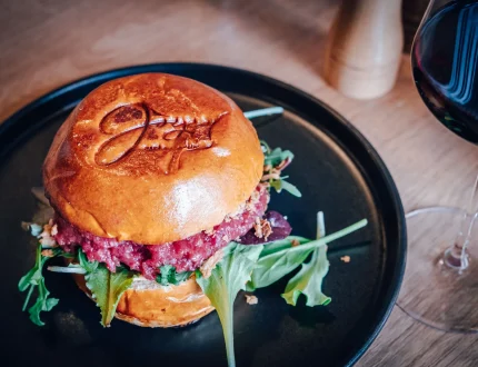 Ein Gourmet-Burger mit Rucola und einer dicken Rinderpastete in einem glänzenden Brioche-Brötchen liegt auf einem schwarzen Teller neben einem Glas Rotwein auf einem Holztisch.