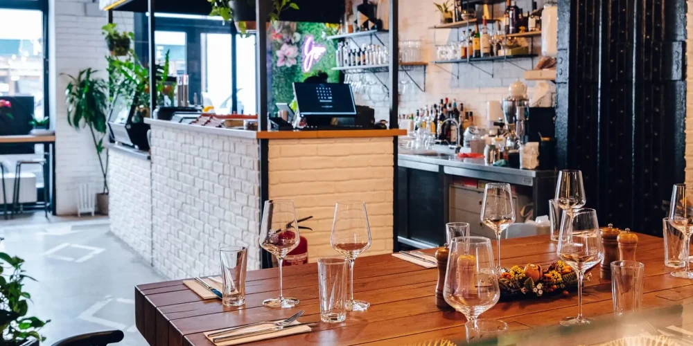 Ein modernes Restaurantinterieur mit einem Holztisch, der mit Gläsern, Besteck und einem dekorativen Mittelstück gedeckt ist. Der Hintergrund zeigt einen Barbereich mit Regalen, Flaschen und Pflanzen, die ein gemütliches und stilvolles Ambiente schaffen.