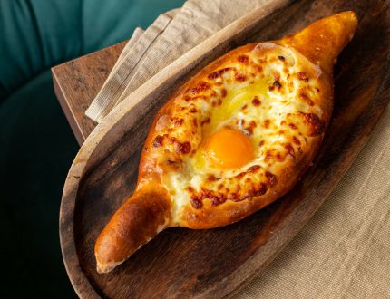 Ein frisch gebackenes Khachapuri, ein schiffchenförmiges Brot, gefüllt mit geschmolzenem Käse und einem Spiegelei, liegt auf einem rustikalen Holzteller auf einer beigen Stoffserviette in einem georgischen Restaurant in Wien.