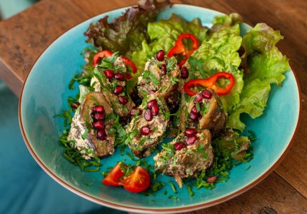 Modi1080_Melanzani-Badrijani Georgisches Restaurant Ein blauer Teller mit Salat, Kirschtomaten, Paprikastreifen und Brötchen, gefüllt mit einer cremigen Masse, garniert mit Granatapfelkernen und frischen Kräutern - ein einladendes Gericht in einem georgischen Restaurant in Wien auf einem Holztisch.