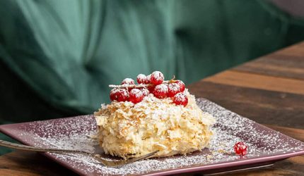 Ein viereckiger Teller mit einem Schichtteigdessert mit roten Beeren und Puderzucker, serviert in einem georgischen Restaurant in Wien, steht auf einem Holztisch mit einem verschwommenen grünen Stuhl im Hintergrund.