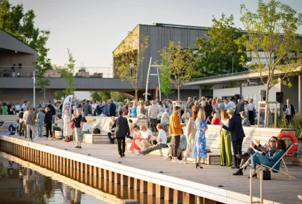Ein lebhaftes Treffen unter freiem Himmel am Wasser, mit Menschen, die auf einer hölzernen Promenade in der Nähe von modernen Gebäuden und Bäumen im warmen Sonnenlicht sitzen und stehen.