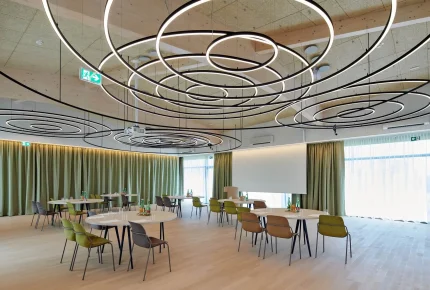 Ein moderner Konferenzraum mit runden Tischen und farbenfrohen Stühlen, großen runden Ringleuchten an der Decke, Vorhängen an den Wänden und einer Projektionsfläche an der Stirnseite. Der Raum ist durch natürliches Licht gut beleuchtet.