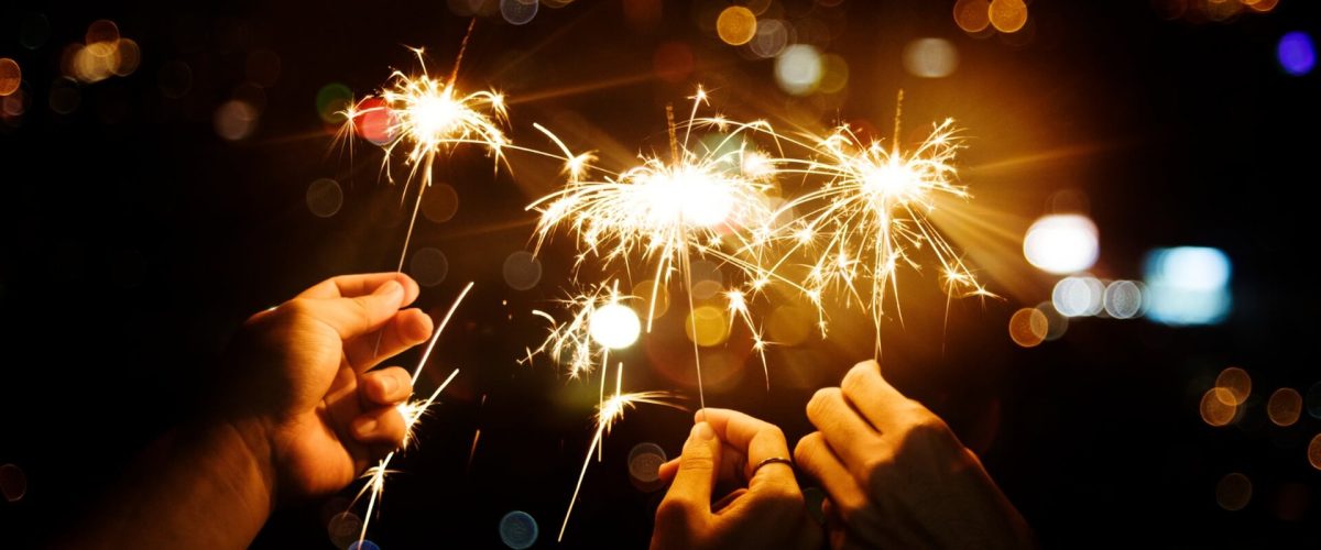 Neujahrsvorsätze_Wo Feiern_Tipps Drei Hände, die Wunderkerzen halten, die in der Nacht hell leuchten, mit einem unscharfen Hintergrund aus Lichtern der Stadt, der den festlichen Geist der Neujahrsvorsätze und der Feierlichkeiten einfängt.