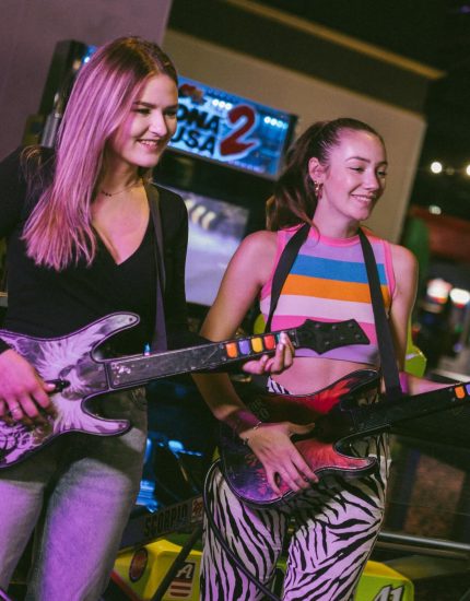 Zwei junge Frauen, die lächelnd vor einem Spielautomaten in der Spielhalle Ocean Park Wien stehen und mit einem Gitarren-Controller spielen. Eine trägt ein schwarzes Oberteil und Jeans, die andere ein bunt gestreiftes Oberteil und eine Hose mit Zebramuster.