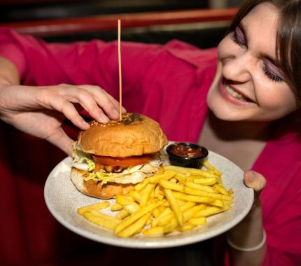 Eine lächelnde Frau in einem rosa Hemd hält einen Teller mit einem Burger und Pommes frites im Ocean Park Wien, mit einem kleinen Becher Soße an der Seite. Sie ist im Begriff, den Burger in die Hand zu nehmen.