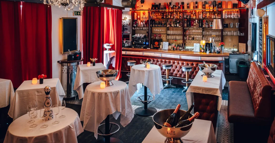 Eine gemütliche Bar im Party Wien mit runden, weiß gedeckten Tischen, auf denen jeweils Eiskübel mit Sektflaschen und Kerzen stehen. Die gut gefüllte Bar, die roten Vorhänge und die warme Beleuchtung schaffen eine einladende Atmosphäre für jede Feier.