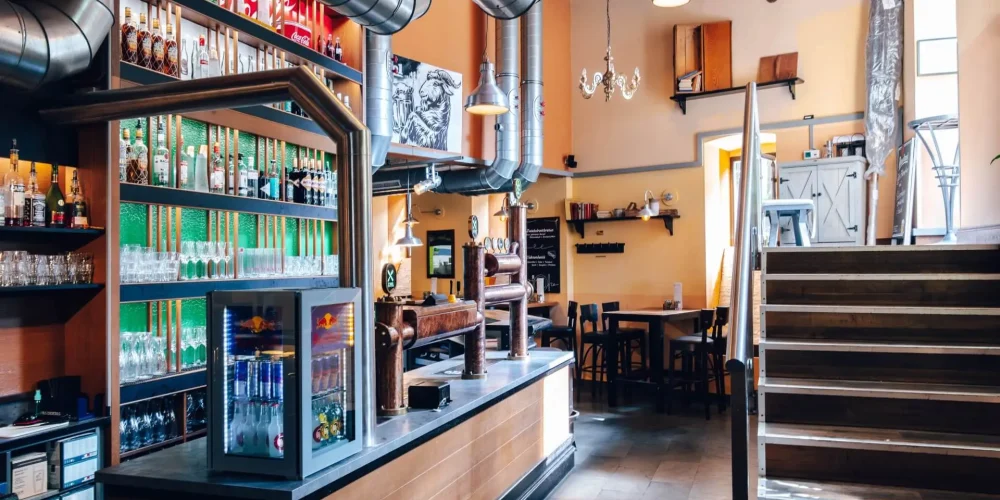 Modernes Barinterieur mit Metall- und Holzdekor, Regalen mit Flaschen, Getränkeautomaten und einem kleinen Kühlschrank. Es gibt Tische und Stühle, Wandkunst, Hängelampen und eine Treppe, die nach oben führt.