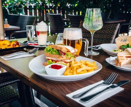 Ein für eine Mahlzeit im Freien gedeckter Tisch mit Tellern mit Sandwiches, Pommes frites mit Ketchup, Salaten, Getränken, darunter Bier und Cocktails, und Besteck auf Servietten. Der Hintergrund zeigt Grünzeug und Korbstühle.
