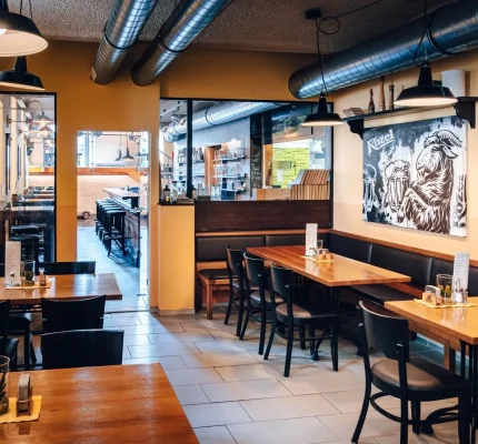 Ein gemütliches, leeres Restaurant mit Holztischen und schwarzen Stühlen, Deckenlüftern im Industriestil, warmen gelben Wänden und einem schwarz-weißen Wandgemälde mit einem Hahn an einer Wand. Speisekarten, Salz- und Pfefferstreuer stehen auf den Tischen.