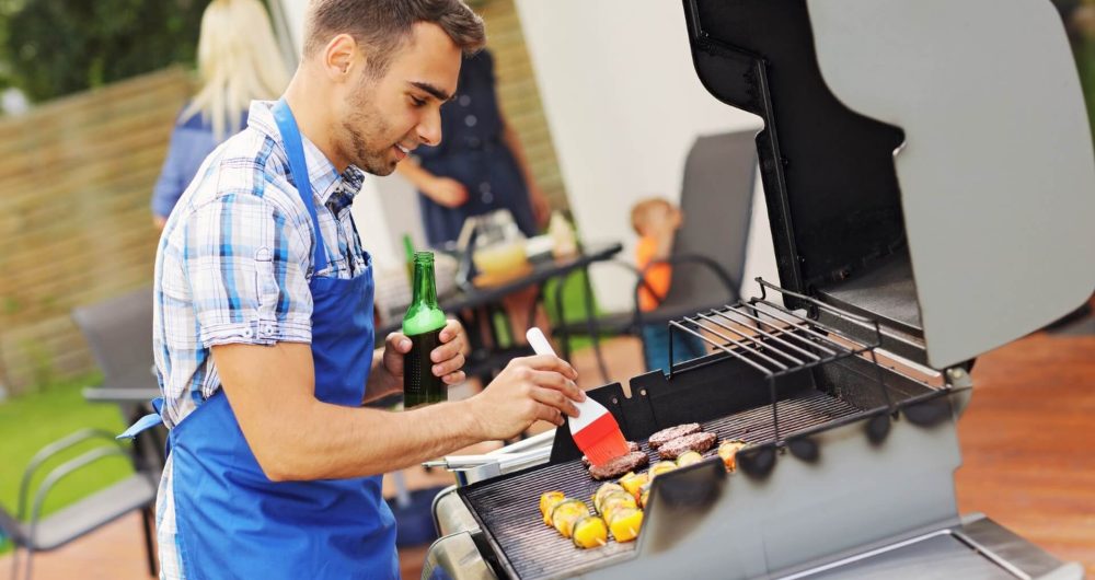 Ein Mann in einer blauen Schürze genießt das Grillen im Freien, grillt Spieße und Burger, während er ein Getränk in der Hand hält und das Essen begießt. Im Hintergrund sieht man andere Personen, darunter ein Kind, die sich unterhalten.
