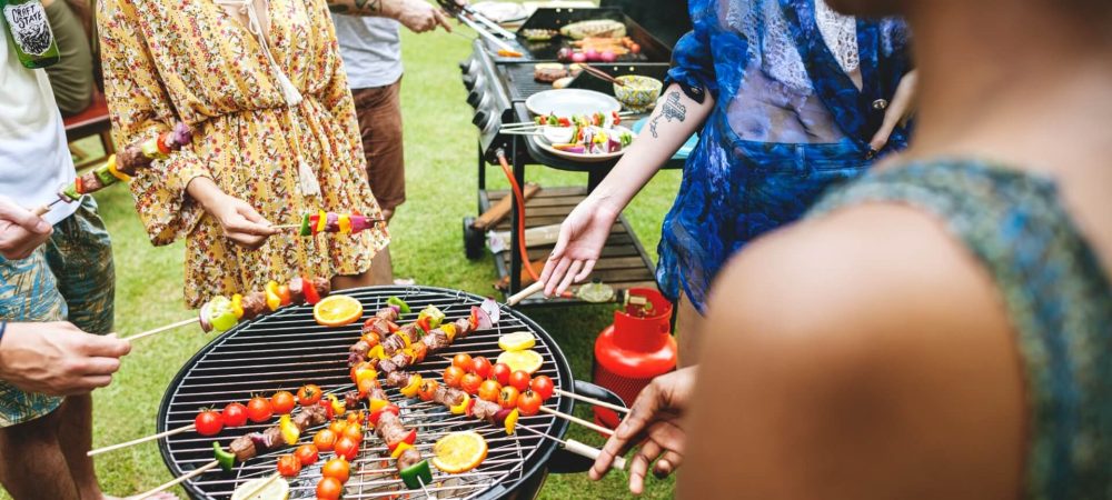 Eine Gruppe von Menschen versammelt sich um einen Grill im Freien und genießt Grillen mit Gemüse- und Fleischspießen. Einige halten Spieße in der Hand, während andere im Hintergrund das Essen zubereiten, bei diesem zwanglosen Barbecue auf grünem Gras.