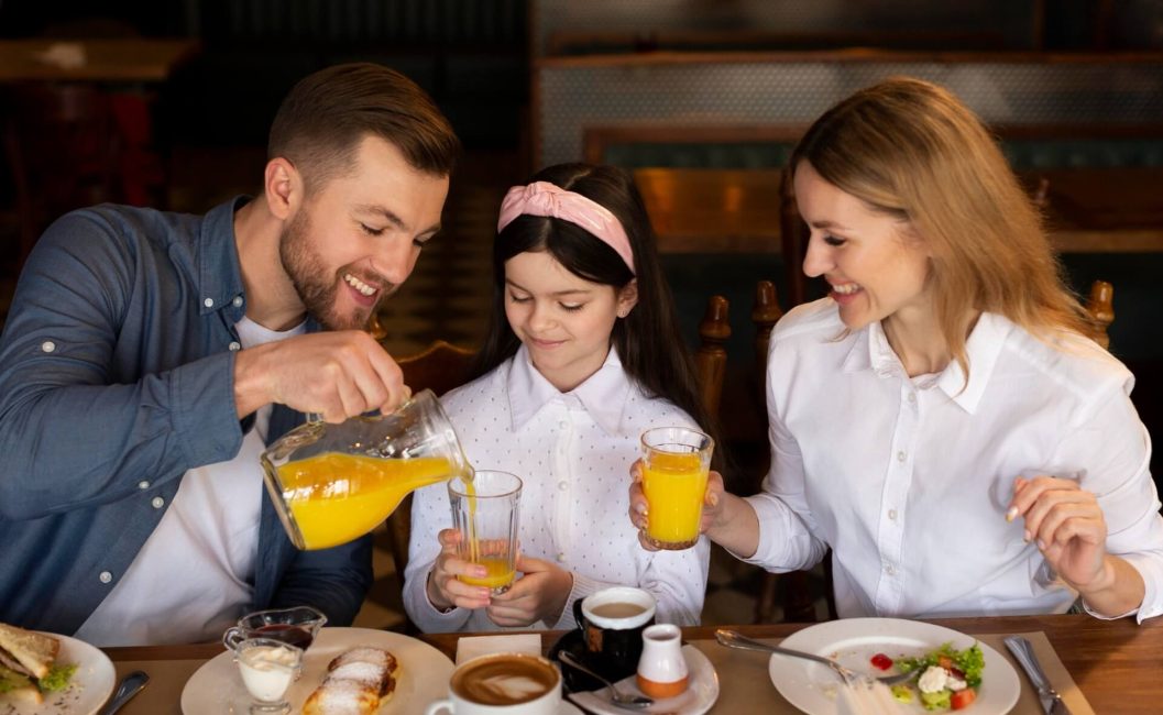 Ein Mann gießt Orangensaft aus einem Krug in das Glas eines Mädchens, während eine Frau lächelt; sie sitzen gemeinsam an einem Tisch und frühstücken mit Gebäck, Kaffee und Saft - sie feiern den Vatertag in Wien in herzlicher Gesellschaft.