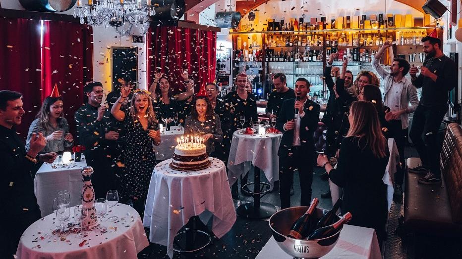 Eine Gruppe von Menschen feiert auf einer Party in einem gemütlichen, gut beleuchteten Restaurant. Konfetti fällt, als eine Frau neben einer Torte mit brennenden Kerzen steht, während andere lächeln, klatschen und Fotos an Tischen mit Getränken und Champagner machen.