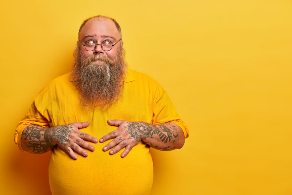 Weihnachtsspeck loswerden, Mann mit Bauchd-man-keeps-hands-big-tummy-stands-thoughtful-pose-has-tattooed-arms-thick-beard-wears-round-glasses-isolated-yellow-wall-blank-space-aside-thinks-doubts (1)