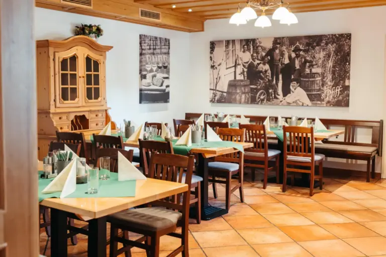 Ein gemütlicher Restaurantbereich mit Holztischen und -stühlen, die für die Gäste gedeckt sind, grünen Tischläufern, Servietten und Gläsern. An den Wänden hängen große Schwarz-Weiß-Fotos und ein Holzschrank. Warme Beleuchtung und ein Fliesenboden sorgen für eine einladende Atmosphäre.