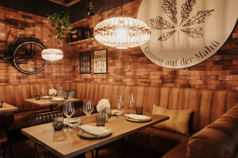 Ein gemütliches Restaurant mit braunen Ledersesseln, Holztischen mit Tellern und Gläsern, Backsteinwänden, warmen Hängelampen, dekorativen Blumen und einem großen Wandbild mit einem Blatt und einem deutschen Text.