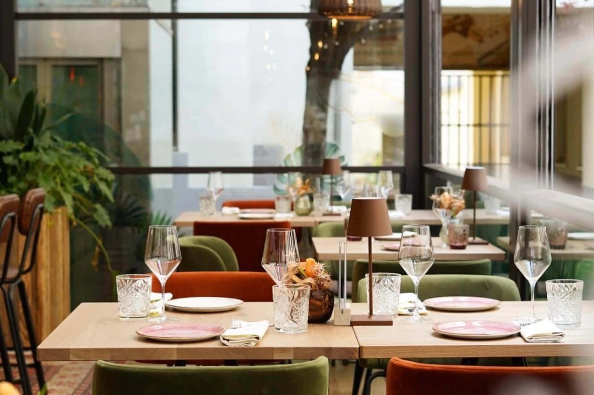 Ein gemütliches, modernes Restaurant mit hübsch gedeckten Tischen, bunten Stühlen, Weingläsern, Tellern, Servietten und kleinen dekorativen Lampen. Große Fenster lassen natürliches Licht herein und bieten einen Blick auf Pflanzen und die Straße draußen.