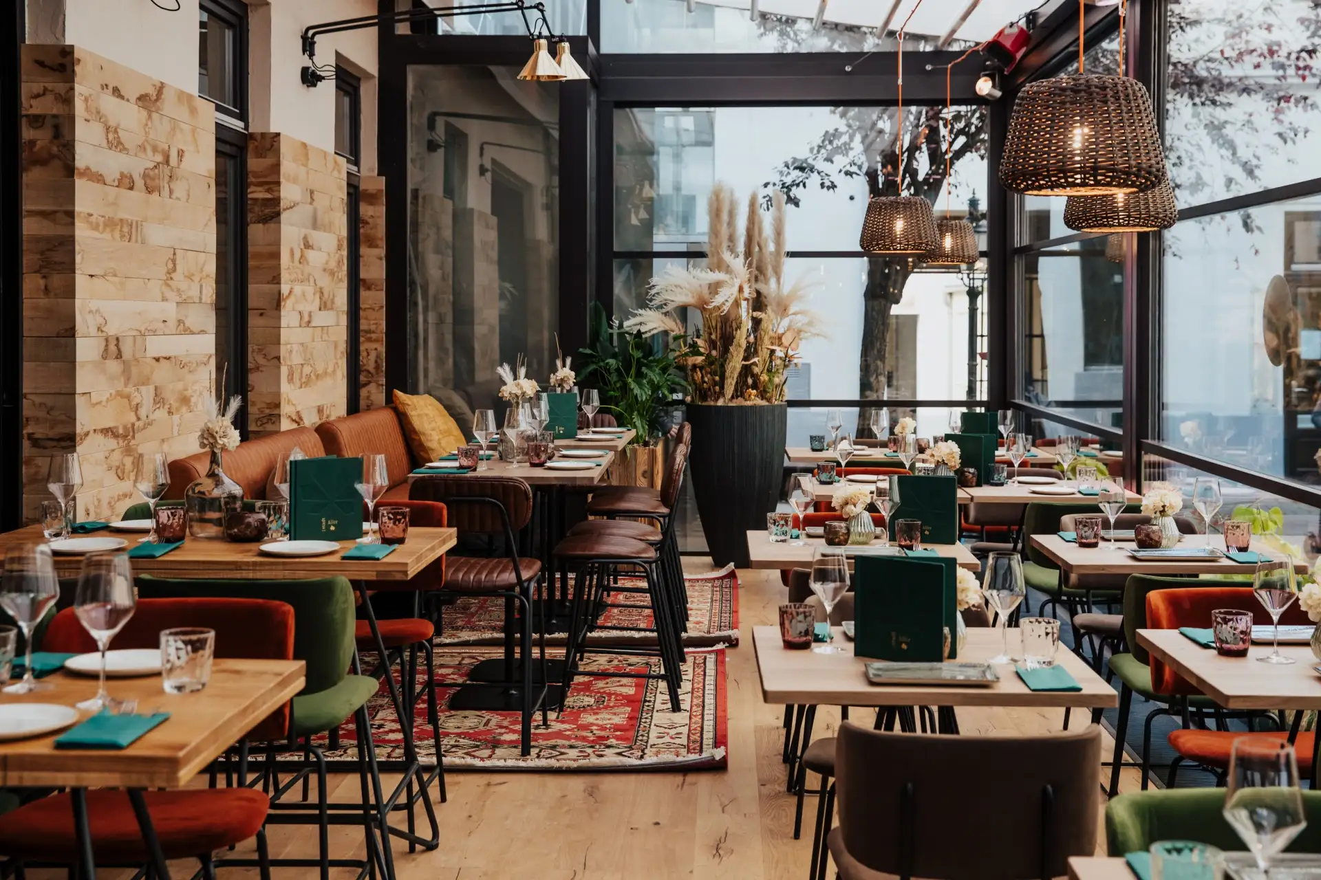 Ein modernes, gemütliches Restaurant mit Holztischen, gepolsterten Stühlen, dekorativen Pflanzen, Hängelampen und großen Fenstern, die natürliches Licht hereinlassen. Die Gedecke und Speisekarten sind ordentlich auf jedem Tisch angeordnet.