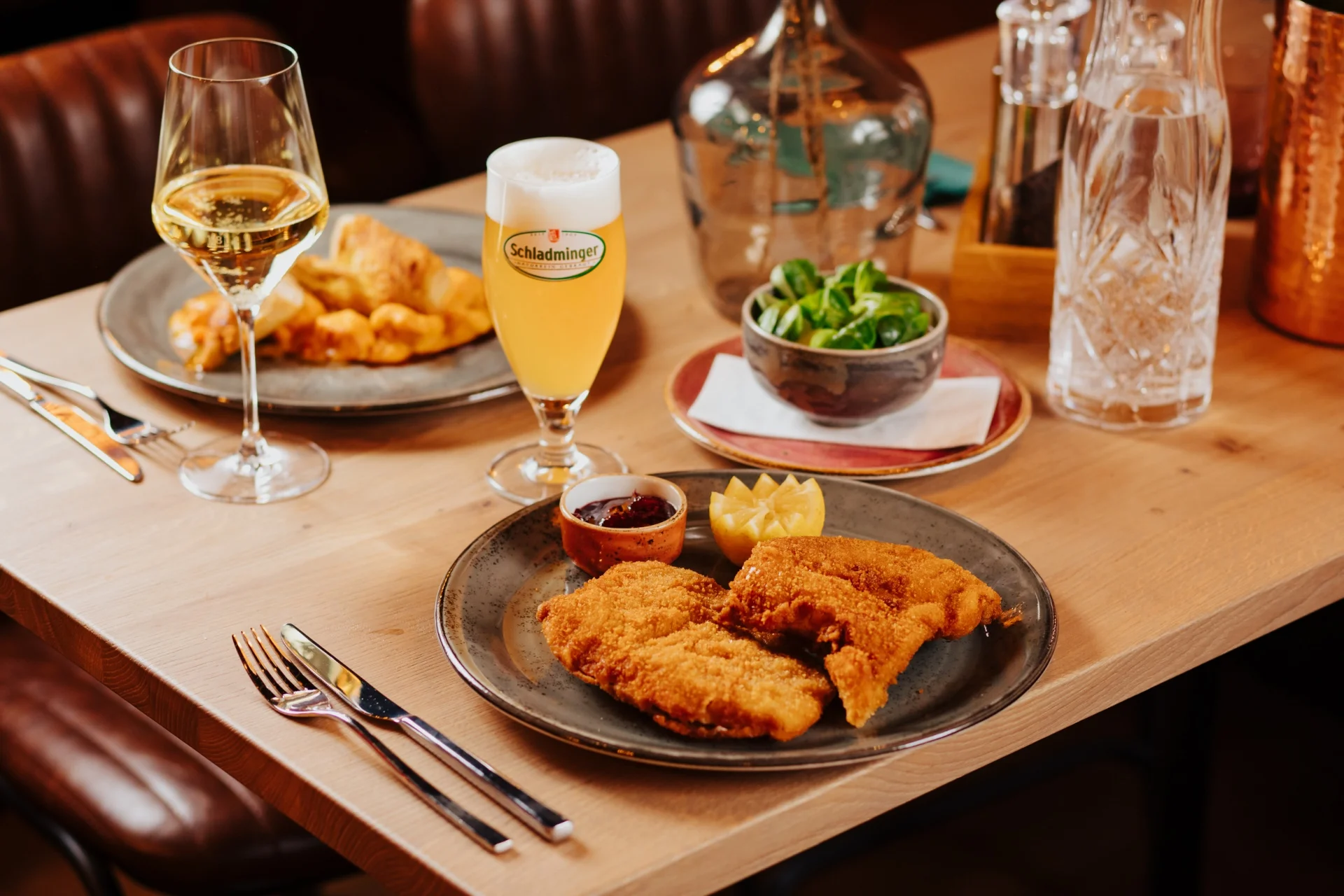 Ein Restauranttisch mit Tellern mit gebratenem Fisch, einer Schüssel mit grünem Salat, einem Glas Weißwein, einem Glas Bier und einer Karaffe mit Wasser, das Besteck ordentlich an der Seite platziert.