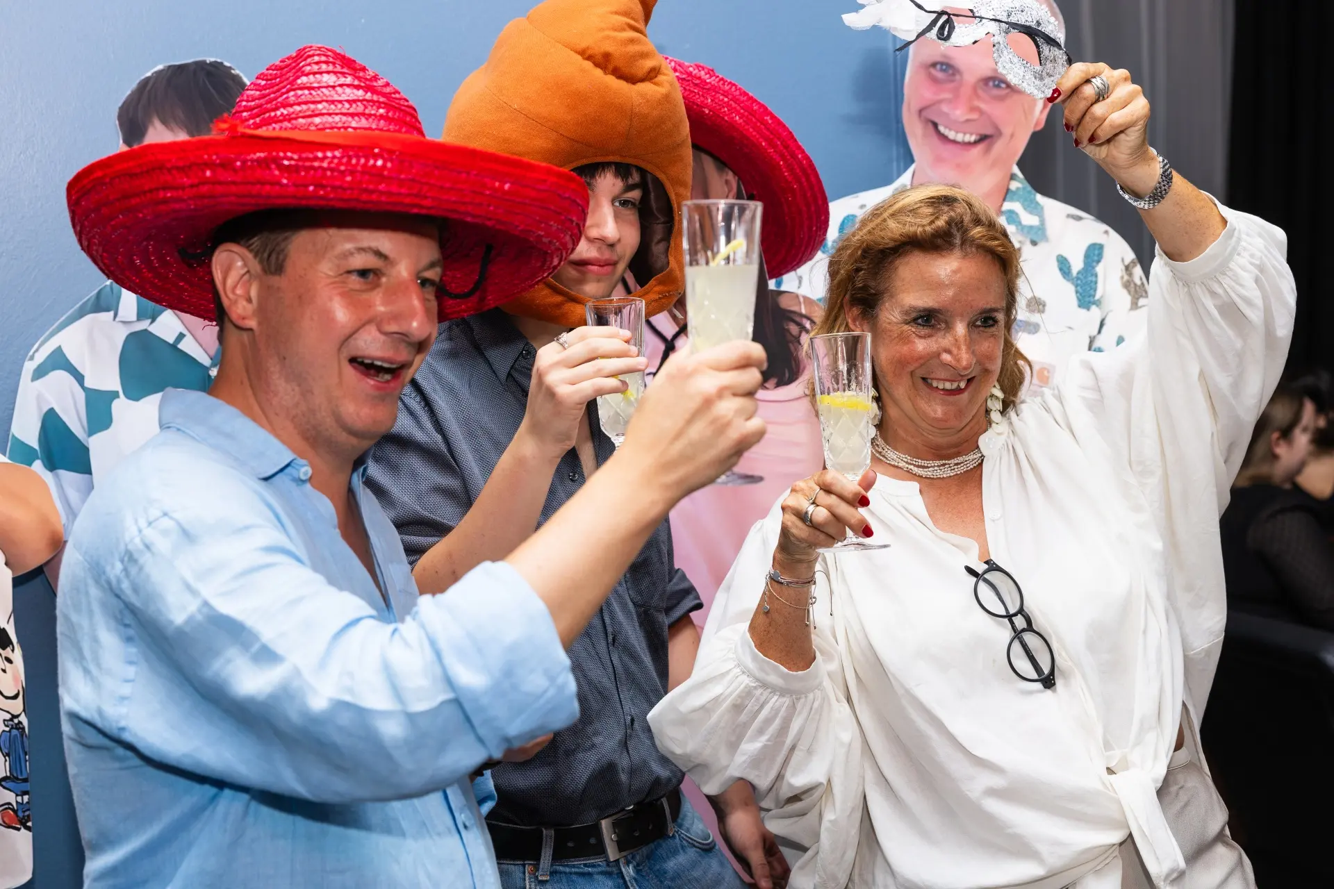 Drei Personen in festlichen Kostümen und mit Hüten halten Gläser hoch und lächeln für ein Foto auf einer Party, mit einer bunten Kulisse im Hintergrund.