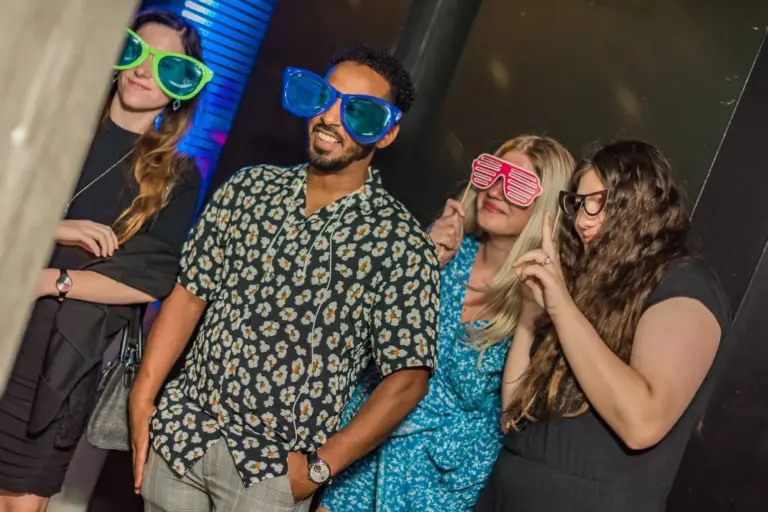Vier Personen posieren zusammen auf einer Party, lächeln und tragen übergroße Scherzbrillen in hellen Farben. Der Hintergrund ist mit gedämpfter Beleuchtung und blauen Akzenten versehen, die eine lustige Atmosphäre schaffen.