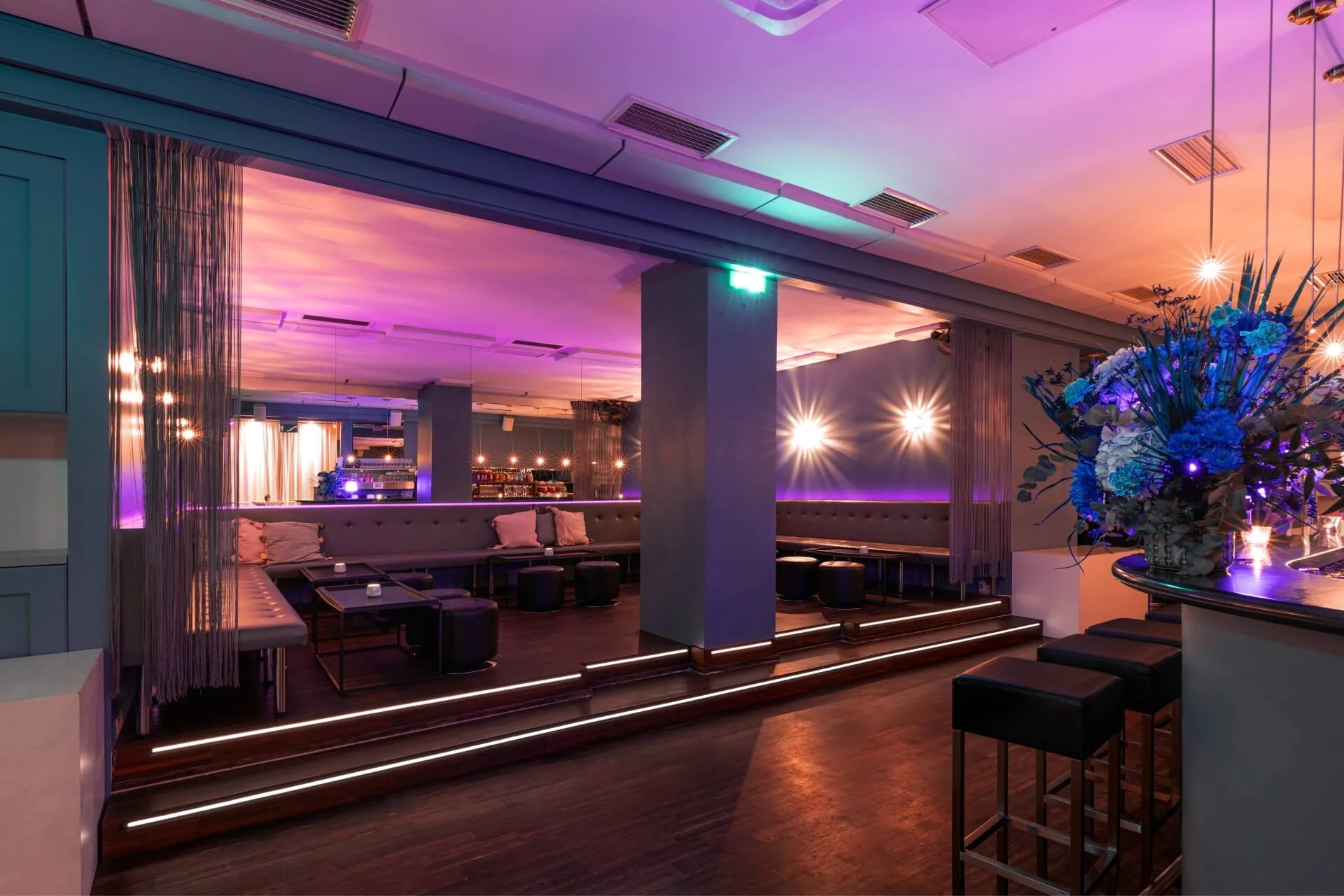 Moderne Lounge mit schummriger, farbenfroher Beleuchtung, grauen Sitzgelegenheiten an den Wänden, kleinen Tischen, hohen Barhockern und Blumenarrangements auf der Bar; die Atmosphäre ist stilvoll und einladend.