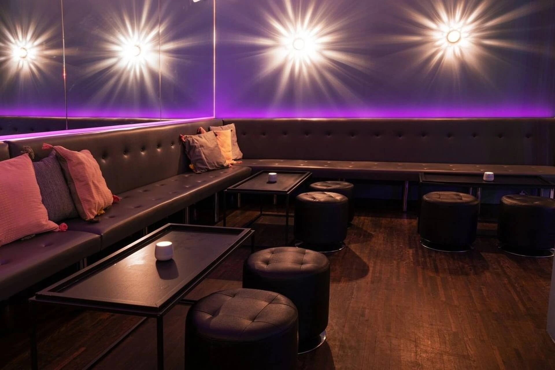 Eine moderne Lounge mit schwarzen, getufteten Sitzmöbeln, niedrigen Tischen, runden schwarzen Hockern und dekorativen Kissen. Der schummrige Raum ist mit Starburst-Wandleuchten und einem lilafarbenen LED-Streifen an der Wand ausgestattet, die ein gemütliches Ambiente schaffen.