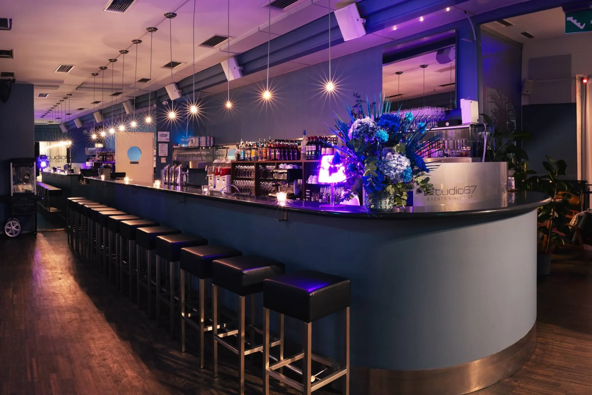 Eine moderne, schwach beleuchtete Bar mit einem langen Tresen, schwarzen Barhockern, hängenden Pendelleuchten, Regalen mit Flaschen, einem Strauß blauer Blumen und sanfter lila Akzentbeleuchtung.