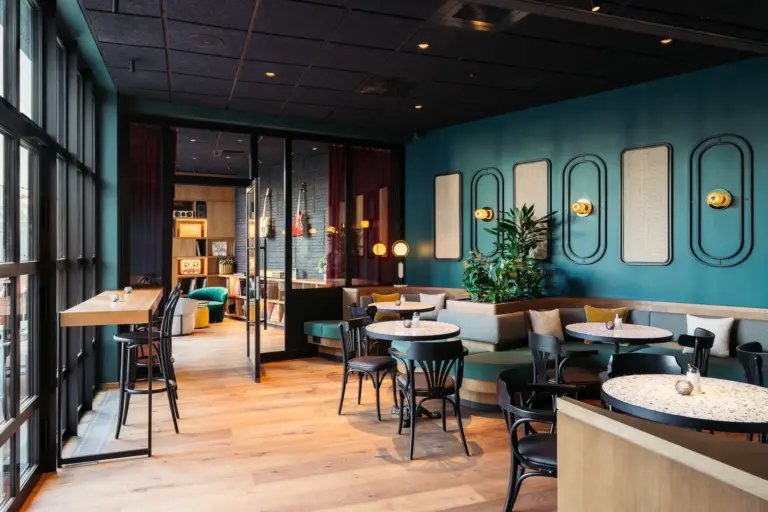 Modernes Café mit bodentiefen Fenstern, türkisfarbenen Wänden, runden Tischen, schwarzen Stühlen, einer Eckbank, Holzfußboden und warmer Beleuchtung. Grünpflanzen und Wandkunst tragen zu einer gemütlichen, einladenden Atmosphäre bei.