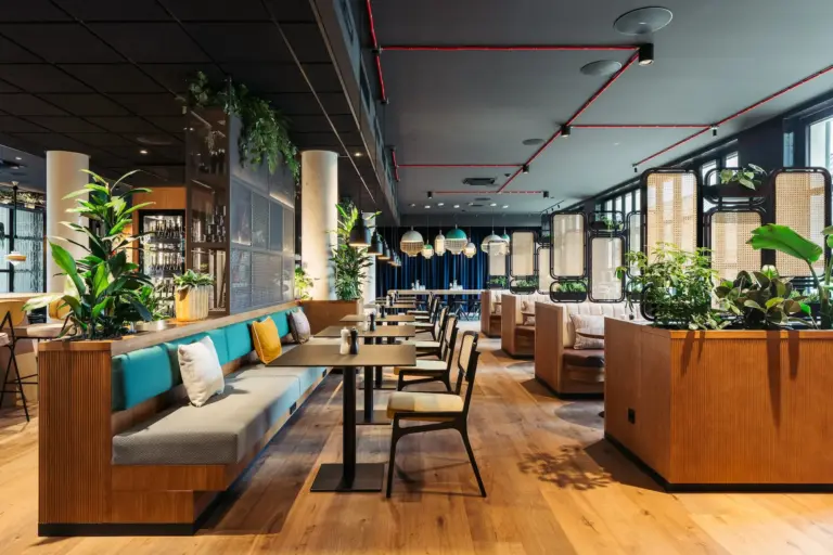 Die moderne Einrichtung des Restaurants mit Holzböden, gepolsterten Sitzbänken, Tischen und Stühlen, großen Fenstern, Hängelampen und vielen Grünpflanzen schafft eine helle und einladende Atmosphäre.