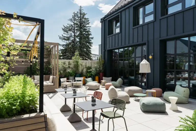 Moderne Außenterrasse mit verschiedenen bunten Stühlen, Tischen, weichen Hockern und Grünpflanzen. Der Raum befindet sich neben einem dunkel getäfelten Gebäude mit großen Fenstern, unter einem sonnigen blauen Himmel. Gemütliche und einladende Atmosphäre.