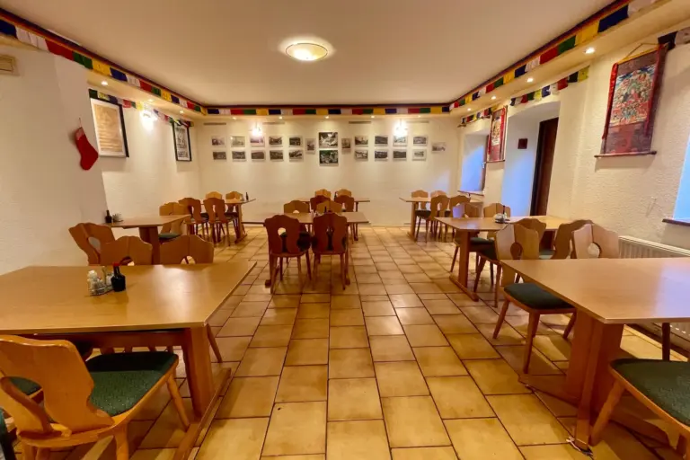 Ein gemütlicher, leerer Speisesaal mit Holztischen und -stühlen, gefliestem Boden, gerahmten Fotos an den Wänden, bunten Bannern an der Decke und einem einzelnen roten Strumpf, der an der linken Wand hängt.