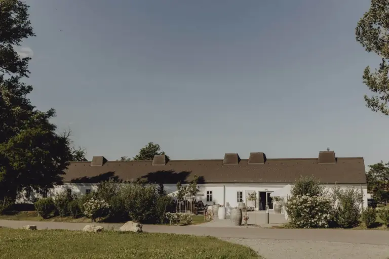 Ein langes, weißes, einstöckiges Gebäude mit einem dunkelbraunen Dach steht unter einem klaren blauen Himmel, umgeben von Grün, Büschen und einem Kiesweg im Vordergrund.